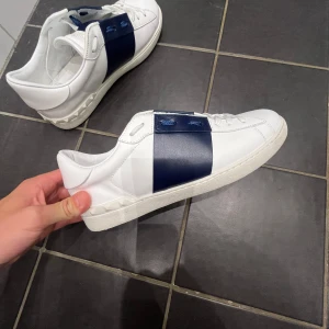 Valentino Garavani Open sneakers vit/blå - Säljer ett par Valentino Garavani Open sneakers i vitt skinn med bred marinblå läderdetalj över snörningen. Snygga platta sulor med mönster och ikoniska nitar på hälen. Rund tå och klassisk siluett, perfekt för dig som gillar exklusiv streetstyle. Nästan helt nya, använde dem 2 gånger och storleken var tyvärr inte helt bra och därför jag säljer dem.Köpte dem själv  för 4399kr av en kompis som hade köpte dem från merch Sweden. Vid snabb affär kan priset diskuteras lite😁🤩