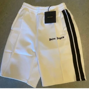 Palm Angels shorts - Palm Angels vita shorts med svarta sidoremsor och broderad logga på framsidan. De har elastisk midja och sportig look, perfekta för dig som gillar streetwear. Materialet känns som polyester och passformen är normal. Snygga detaljer med kontrastfärgade ränder.