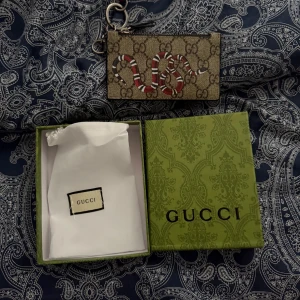 Gucci plånbok med orm och monogram - Snygg plånbok från Gucci med klassiskt GG-monogram och en cool röd, vit och svart orm på framsidan. Plånboken har metallkedja med karbinhake och levereras i original ask och dustbag. Perfekt för kort och smågrejer.
