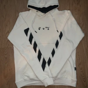Off-White vit hoodie med svarta detaljer - Säljer en snygg vit hoodie från Off-White i storlek M. Tröjan har svarta diagonala ränder på framsidan, broderad Off-White-logga och coola snören vid huvan. Insidan av huvan är svart för extra kontrast. Perfekt för dig som gillar streetwear och vill sticka ut.