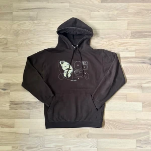 Backstory hoodie - Brun hoodie från Back Story i storlek S med ett unikt tryck på bröstet som visar en fjäril och geometriska former. Tröjan har huva med snörning och en klassisk känguruficka framtill. Materialet är mjuk bomull blandat med polyester, perfekt för chill dagar.