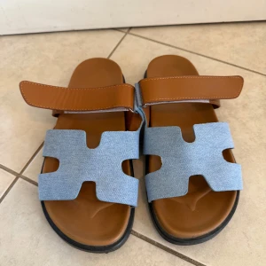 Bruna och ljusblå sandaler strl 41 - Säljer ett par sandaler med cool design i brunt och ljusblått. De har breda remmar i ljusblå textil och en justerbar brun rem i skinn över foten. Svart platt sula med mönstrad undersida för extra grepp. Perfekt för sommardagar och chill häng.