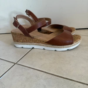 Bruna kilklackssandaler från Anna Field - Snygga bruna sandaler från Anna Field med kilklack i kork och detaljerad flätad kant. Remmarna är i brunt syntetmaterial med justerbart spänne. Sandalerna har öppen tå och en vit platt sula som ger en fräsch kontrast. Perfekta för sommarens alla dagar! Knappt använda då dom var för små. Obs, det är en spricka i det nedre vita materialet på höger sko (se sista bilden)