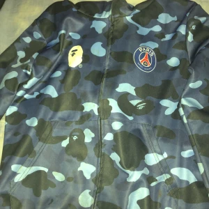 BAPE x PSG blå camo vindjacka - Snygg vindjacka från BAPE x PSG med blått camo-mönster och feta tryck som hajansikte på ryggen, BAPE-logga och PSG-märke på bröstet. Jackan har dragkedja framtill och är tillverkad i polyester. Perfekt för dig som gillar streetwear och vill sticka ut.