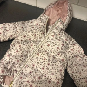 Blommig pufferjacka med huva - Säljer en söt pufferjacka med blommigt mönster i rosa och lila toner på vit bakgrund. Jackan har huva, dragkedja och quiltad design. Insidan är ljusrosa och materialet känns lätt och mjukt, perfekt för kyliga dagar. Passar dig som gillar romantisk och chill stil.