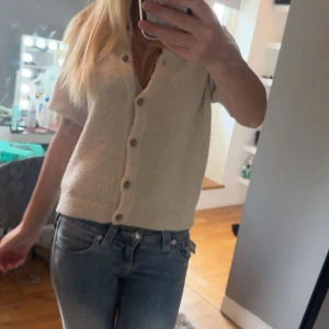 Beige kortärmad kofta med knappar - Säljer en beige kortärmad kofta med snygga knappar framtill. Tröjan har ribbad kant nedtill och vid ärmslut, vilket ger en schysst passform. Perfekt att slänga på över ett linne eller t-shirt för en chill look.