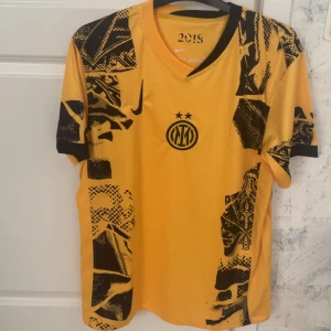 Gul Inter Milan fotbollströja Nike - Snygg gul Inter Milan fotbollströja från Nike med svart grafiskt mönster och klubbens logga på bröstet. Tröjan har korta ärmar, V-ringning och är tillverkad i lätt, ventilerande material som passar perfekt på planen. Limited edition 2018. 