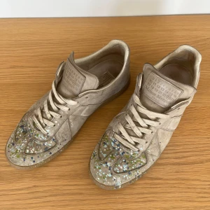 Maison Margiela  - Maison Margiela sneakers i beige mocka med coola färgstänk i blått, grönt och vitt över tån. Klassisk rund tå, snörning och platt sula. Märkets ikoniska etikett på plösen. Perfekt för dig som gillar unik design och streetstyle.