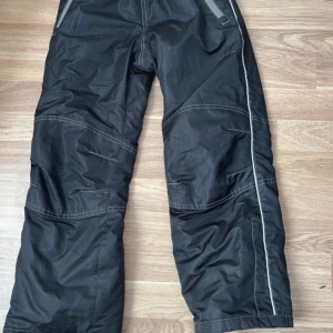 Svarta täckbyxor från Ellos, stl 134 - Svarta täckbyxor från Ellos med elastisk midja och dragkedjeförsedda fickor. Byxorna har synliga sömmar och är tillverkade i slitstarkt syntetmaterial, perfekta för kalla dagar. Snyggt sportig look med praktiska detaljer.