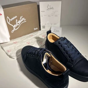 Mörkblå sneakers från Christian Louboutin - Snygga mörkblå sneakers från Christian Louboutin i mocka med klassisk röd sula och broderad logga på hälen. Skorna har rund tå, platt sula och snörning framtill. Insidan är beige och ger en lyxig känsla. Perfekta för dig som gillar exklusiv streetstyle.