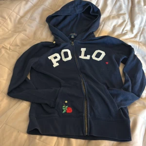 Marinblå hoodie från Polo Ralph Lauren - Snygg marinblå hoodie från Polo Ralph Lauren med vit POLO-text på bröstet och en liten röd broderad ros vid fickan. Tröjan har huva, dragkedja och långa ärmar. Priset kan diskuteras