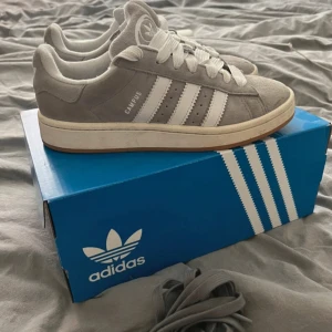 Grå adidas Campus sneakers, storlek 38 - Säljer ett par grå adidas Campus sneakers med klassiska vita ränder och logga på hälen. Skorna har rund tå, platt sula och mjuk mocka med vita snören. De är använda max 4 gånger !