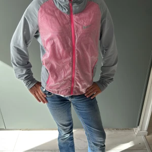 Nike Golf vindjacka XS rosa/grå - Nike Golf Tour Performance vindjacka i Dri-FIT material, perfekt för aktiva dagar. Jackan är rosa med grå ärmar och detaljer, har ett diskret rutmönster och hel dragkedja framtill. Sportig look med långärmad passform och meshpaneler för extra ventilation.