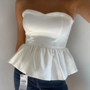Vit axelbandslös peplum topp - Supersnygg glansig vit topp med peplumvolang nedtill.🤗 Toppen har även en inbyggd ”BH”. Den har vissa skavanker som alla syns på bilderna. 