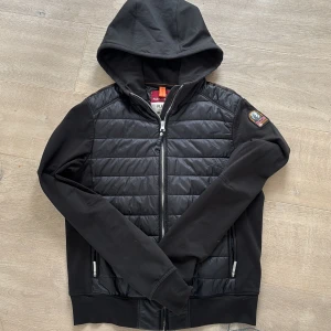 Svart Parajumpers puffer hoodie - Svart pufferjacka från Parajumpers med huva och dragkedja. Jackan har quiltad framsida i syntetmaterial och mjuka fleeceärmar. Märkets patch på ena ärmen ger en cool touch. Perfekt för dig som gillar streetstyle och vill ha något som sticker ut.