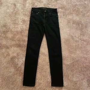 Svarta Nudie Jeans Hightop Tilde - Snygga svarta jeans från Nudie Jeans, modellen Hightop Tilde i färgen Raven Black. De har hög midja, smal passform och klassiska fem fickor. Materialet är 92% bomull, 6% polyester och 2% elastan för extra stretch. Diskret Nudie Jeans-logga på bakfickan och svart läderpatch.