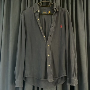 Blå skjorta från Polo Ralph Lauren - Svart långärmad skjorta från Polo Ralph Lauren med klassisk krage och knappar framtill. Broderad röd logga på bröstet ger en snygg kontrast. Skjortan har en loose passform och är tillverkad i mjuk bomull som känns skön mot huden.