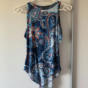 Blommig bodytopp från Boohoo - Snygg bodeytopp från Boohoo med färgstarkt blommigt mönster i marinblått, vitt och orange. Toppen har rund hals och asymmetrisk nederkant som ger en cool vibe. Perfekt för dig som gillar boho-stil och vill sticka ut med färg och mönster.