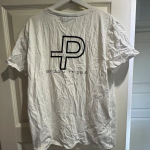 Vit t-shirt från Pelle P med tryck - Vit t-shirt från Pelle P med svart logotyptryck på ryggen och ärmen. Klassisk rund hals och korta ärmar. Tillverkad i mjuk bomull som är skön mot huden. Snyggt minimalistisk design med koordinater under loggan på ryggen.