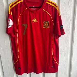 Spanien David Villa #7 EM 2008 tröja - Säljer en officiell röd Spanien fotbollströja från EM 2008 med David Villa och nummer 7 i guld på ryggen. Adidas-logga och landslagsemblem på bröstet, gula detaljer och ränder på axlarna. Kortärmad och tillverkad i lätt polyester, perfekt för match eller samling.