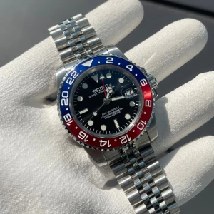 Seiko GMT-Master II Pepsi mod - Helt nybyggd Seiko GMT-Master 2 ”Pepsi” Mod  Specifikationer: Storlek: 40mm Urverk: Nh34 (Automatiskt med GMT Funktion) Material: 904L Rostfritt Stål Glas: Sapphire Glas med Förstoring vid Datum Boett: Pepsi Färgerna på Bezel Tavla: Svart med Datum Armband: Viklås och Justerbart (Jubilee) Krona: Screw Down Vattentålighet: 30 Meter  En klassiker! Pepsin är en av dem mest välkända klockorna på marknaden. Med denna klockan kommer du få må