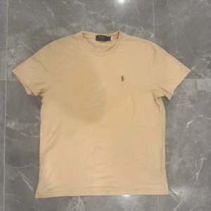 Beige t-shirt från Polo Ralph Lauren - Snygg beige t-shirt från Polo Ralph Lauren i klassisk modell med rund hals och korta ärmar. T-shirten har den ikoniska broderade loggan på bröstet och är gjord i mjuk bomull. Perfekt för en chill och stilren look.