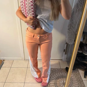 Persiko/coral jeans  - Säljer ett par unika persikofärgade bootcut byxor med breda vita slitsar längs utsidan av benen. Byxorna har klassisk femficksdesign, knapp och dragkedja fram. Kilen är påsydd med maskin och försurning små dragningar i tyget är jeansen i mycket bra skick! 
