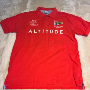 Röd pikétröja från 8848 Altitude - Snygg röd pikétröja från 8848 Altitude med vit text och broderad svensk flagga på bröstet. Klassisk krage med knappslå och korta ärmar. Materialet är mjuk bomull som känns skönt mot huden. Perfekt för dig som gillar sportig och clean stil.