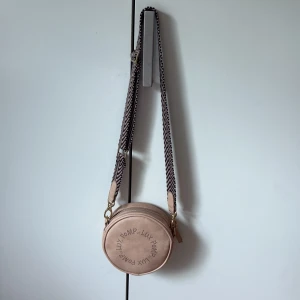 Beige rund axelväska från Pomp de Lux - Säljer en beige rund axelväska från Pomp de Lux med coolt perforerat logomönster på framsidan. Väskan har en bred, svartvit randig axelrem och dragkedja upptill. Materialet är skinnimitation och insidan är fodrad. Perfekt storlek för mobilen och småsaker.
