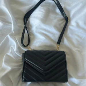 Svart quiltad axelväska med avtagbar axelrem - Snygg svart axelväska med quiltad yta i chevronmönster och guldfärgade metalldetaljer. Väskan har en avtagbar och justerbar axelrem, perfekt för att bära crossbody. Stilren design med lock och magnetstängning