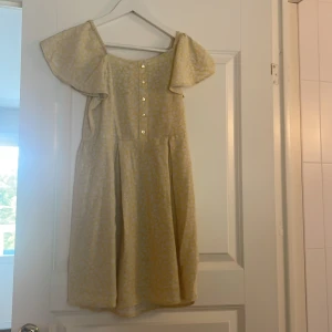 Blommig ljusgul klänning Kappahl - Supersöt ljusgul kortklänning från Kappahl med vitt blommigt mönster. Klänningen har offshoulder-ärmar, knappar framtill och smock i ryggen för skön passform. Perfekt för dig som gillar romantisk och somrig stil!