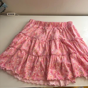 Blommig volangkjol med inbyggda shorts  - Supersöt kort kjol från Lindex med rosa botten och små blommor i vitt och gult. Kjolen har flera volanger och ett elastiskt midjeband för skön passform. Perfekt för dig som gillar färg och mönster! Materialet är mjukt och lätt bomull.