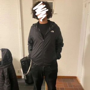 Svart vindjacka från The North Face - Svart vindjacka från The North Face med huva och dragkedja framtill. Jackan har en clean look med logga på bröstet och är tillverkad i lätt polyester. Perfekt för dig som gillar streetstyle och vill ha något enkelt men snyggt.