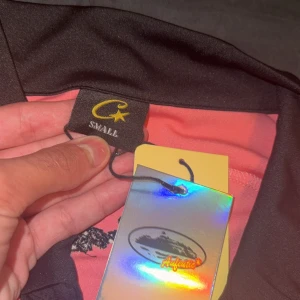 Rosa pikétröja med svart krage - Säljer en rosa pikétröja från Authentic i storlek small. Tröjan har svart krage och svarta detaljer längs ärmar och sidor, samt ett tryckt motiv på bröstet. Materialet känns som polyester och är lätt och skönt. Perfekt för dig som gillar sportig stil.