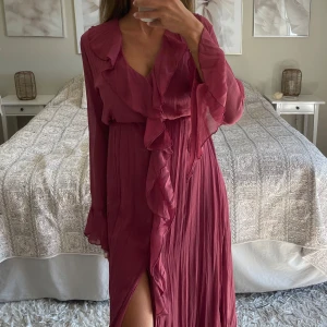 Långklänning med volanger från Zara - Supersnygg långklänning från Zara med v-ringning och volangdetaljer längs framstycket och ärmsluten. Klänningen har transparenta, långa ärmar och ett flowigt, plisserat tyg som ger en romantisk vibe. Perfekt för dig som gillar boho och vill sticka ut.