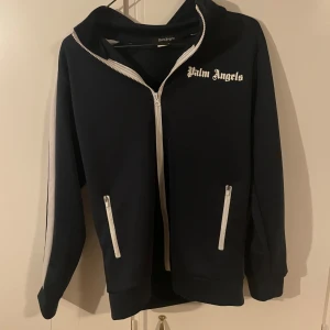 Palm Angels svart bomberjacka - Marinblå  bomberjacka från Palm Angels med vita detaljer och dragkedjor. Jackan har vita ränder längs ärmarna, ståkrage och logotypen broderad på bröstet. Tillverkad i mjukt syntetmaterial, perfekt för en streetwear-look.