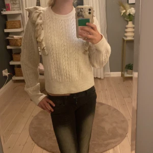 Vit stickad tröja med volang från Lindex - Supersöt vit stickad tröja från Lindex med snyggt kabelmönster och volangdetaljer på ena axeln. Rund hals och långa ärmar, perfekt för dig som gillar en lite extra touch på din outfit. Passar dig som vill ha en mysig men ändå stylish look.