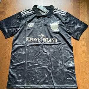 Adidas x Stone Island svart fotbollströja L - Snygg svart fotbollströja i samarbete mellan Adidas och Stone Island. Tröjan har korta ärmar, klassisk krage och silverfärgade detaljer som loggor och ränder på axlarna. Tillverkad i lätt och ventilerande material med diskret mönster över hela tyget.