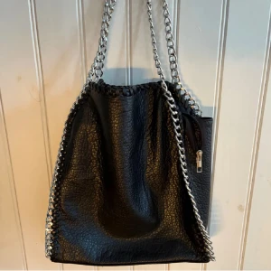 Svart axelväska med kedjedetaljer - Cool svart axelväska från Stella McCartney  i veganskt skinn med grova silverfärgade kedjor längs kanterna och som axelrem. Väskan har dragkedjeficka på utsidan och flera fack inuti. Perfekt för dig som gillar edgy och trendig stil. Kan släppa den för billigare pris vid snabb affär. Väskan är helt oanvänd.