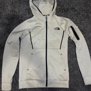 Beige hoodiejacka från The North Face - Snygg beige dress från The North Face med svart logga på bröstet och ryggen. Jackan har hel dragkedja, huva och svarta detaljer längs sömmarna. Praktiska fickor framtill och en extra ficka med dragkedja på ärmen. Perfekt för chill eller träning. Säljer den för 1599. Köpte den för 2,5. säljer den då jag inte har så mycket användning för den och vuxit ur den.