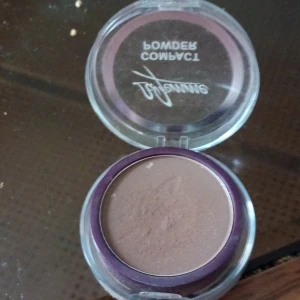 Brun kompakt bronzer - Snygg brun kompakt bronzer i rund plastförpackning med lila kant. Perfekt för att ge ansiktet en varm och naturlig glow. Smidig att ta med i väskan och enkel att applicera med borste.
