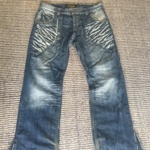 Fishbone blå jeans med detaljer - Säljer ett par blå jeans från Fishbone med coola slitningar och unika mönstrade detaljer på framsidan. Jeansen har custom flarade ben och klassisk femficksdesign. Materialet är robust denim och färgen är snyggt tvättad med ljusare partier.