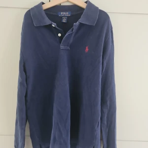 Marinblå pikétröja från Ralph Lauren - Klassisk marinblå långärmad pikétröja från Polo Ralph Lauren med röd broderad logga på bröstet. Tröjan har krage, knappar vid halsen och är i bomull. Perfekt för en stilren och avslappnad look.