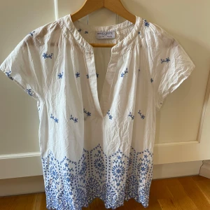 Vit blus med blå broderier Holly & Whyte - Superfin vit blus från Holly & Whyte med blå broderade detaljer och v-ringning. Blusen har korta ärmar, avslappnad passform och vackert mönster längs nederkanten. Perfekt för en fräsch och somrig look!