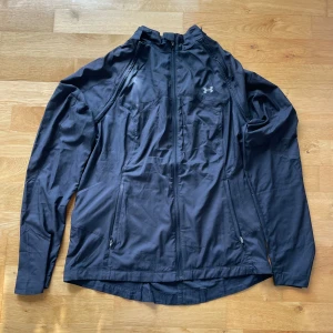 Svart träningsjacka från Under Armour - Svart träningsjacka från Under Armour i 100% polyester. Jackan har hel dragkedja framtill, två fickor med dragkedja och diskret logga på bröstet. Lätt och smidig modell med lång ärm, perfekt för aktiva dagar utomhus.