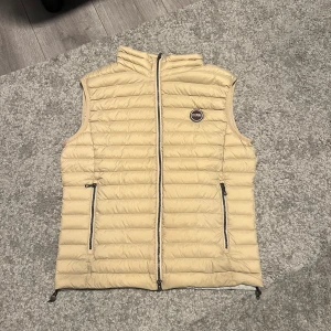 Beige dunväst från Colmar - Snygg beige dunväst från Colmar med quiltad design och två dragkedjefickor framtill. Västen har hög krage och logotyp på bröstet. Perfekt att slänga över en hoodie eller tröja för extra värme. Lätt och smidig att bära, stängs med dragkedja hela vägen.