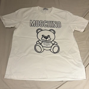 Vit Moschino t-shirt med björntryck - Vit t-shirt från Moschino med stort svart björntryck och texten 'This is not a Moschino toy' på framsidan. Klassisk rund hals och korta ärmar. Skön och mjuk bomull som passar perfekt till jeans eller shorts.