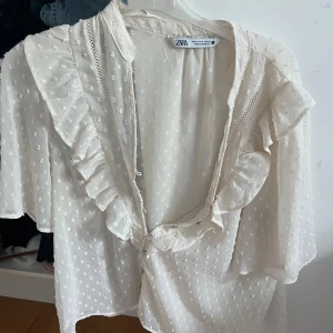 Vit blus med volang  - Supersöt vit blus från Zara med diskret prickigt mönster, volangdetaljer längs v-ringningen och knappar framtill. Blusen har korta, luftiga ärmar och är gjord i ett lätt, transparent tyg som känns riktigt fräscht och somrigt. Storlek S knappt använd, helt ny i skicket 💕💕
