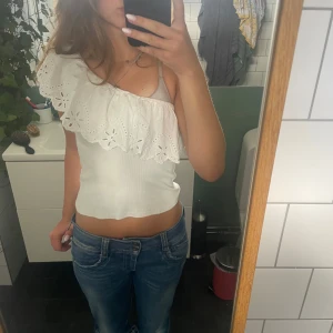 Vit oneshoulder topp med volang Zara - Superfin vit oneshoulder topp från Zara med bred broderad volang och smal axelrem. Toppen är ribbad i tyget och volangen har blommiga broderier som ger en söt vibe. Perfekt till jeans eller kjol för en fräsch look.