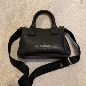 Svart handväska från Valentino - Snygg svart handväska från Valentino med croco-mönstrad yta och guldfärgad logga framtill. Väskan har både handtag och en bred, avtagbar axelrem med justerbar längd. Materialet är skinn och den stängs med dragkedja upptill. Perfekt storlek för det viktigaste.
